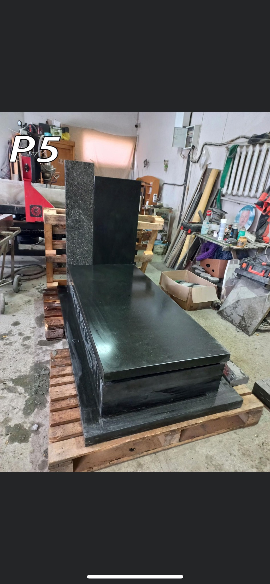 Ciemny, częściowo zmontowany nagrobek z granitu na palecie w warsztacie kamieniarskim; widoczny sprzęt i narzędzia w tle.