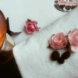 Aromatyczny olejek w szklanym kominku z pływającą różą, obok ręcznik frotte z trzema pąkami róż i trzema czekoladkami.