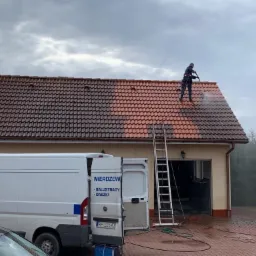 Pracownik w kombinezonie czyści dach z czerwonej dachówki za pomocą myjki ciśnieniowej. Część dachu jest już umyta, kontrastując z brudną resztą. Obok stoi drabina i biały samochód dostawczy...