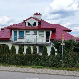 Dom jednorodzinny z nowym, czerwonym dachem i białą elewacją, otoczony zielenią, widoczny balkon z metalową balustradą, antena satelitarna na dachu.