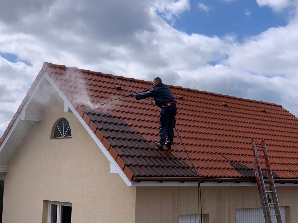 Pracownik w kombinezonie czyści dach z czerwonej dachówki za pomocą myjki ciśnieniowej, widoczna drabina oparta o ścianę budynku z kremową elewacją i okrągłym oknem na poddaszu, błękitne niebo...