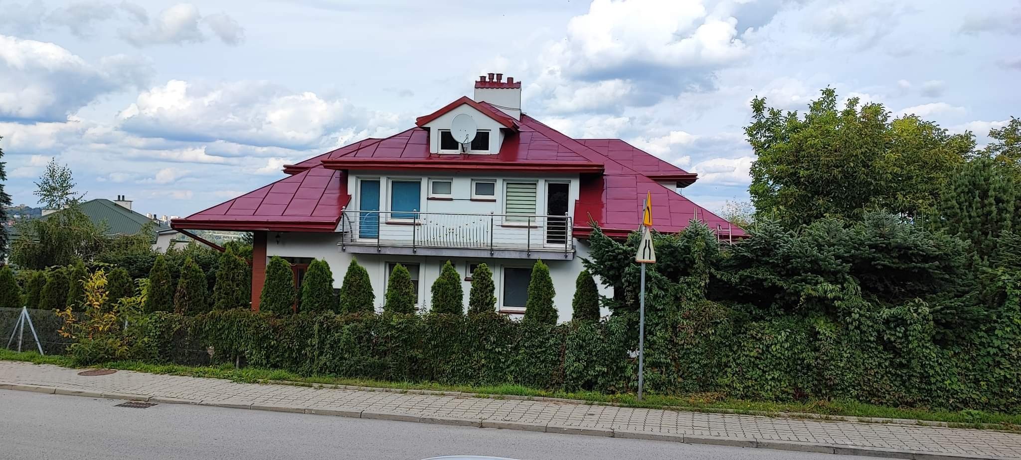 Dom jednorodzinny z nowym, czerwonym dachem i białą elewacją, otoczony zielenią, widoczny balkon z metalową balustradą, antena satelitarna na dachu.