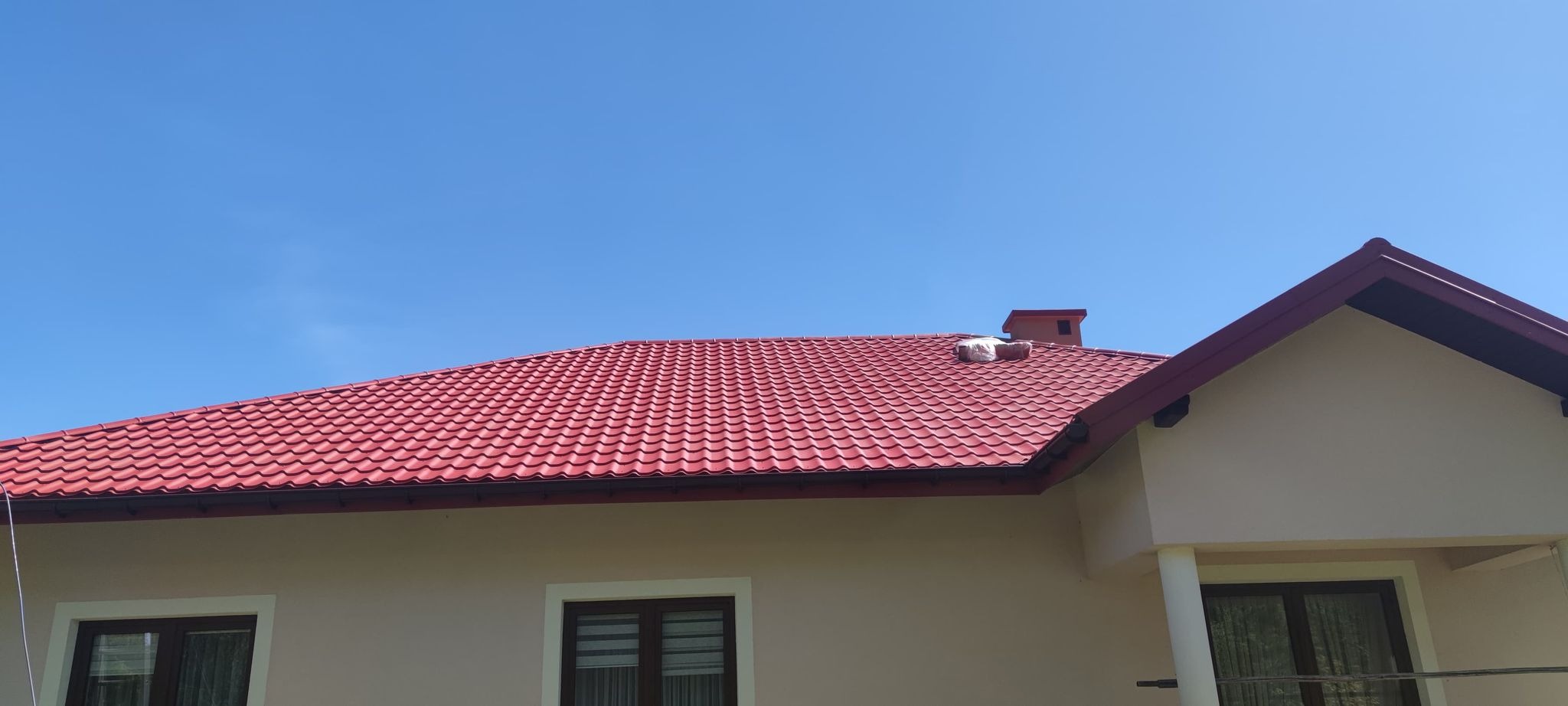 Czerwony dach z blachodachówki na jasnym budynku, fragment komina zabezpieczony folią, błękitne niebo w tle.