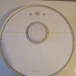 Biały, okrągły robot sprzątający Roborock, leżący na jasnym blacie, widok z góry, pokazujący przyciski i logo.