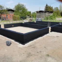 Zabezpieczone, pomalowane na czarno bloczki fundamentowe z widoczną izolacją przeciwwilgociową, przygotowane do dalszych prac budowlanych na działce z zabudowaniami gospodarczymi w tle.