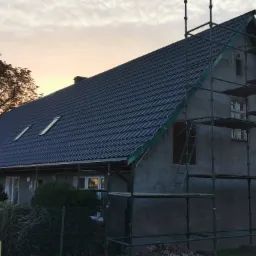 Dwukondygnacyjny budynek z ciemną dachówką w trakcie prac elewacyjnych, otoczony rusztowaniem, widoczny o zmierzchu.