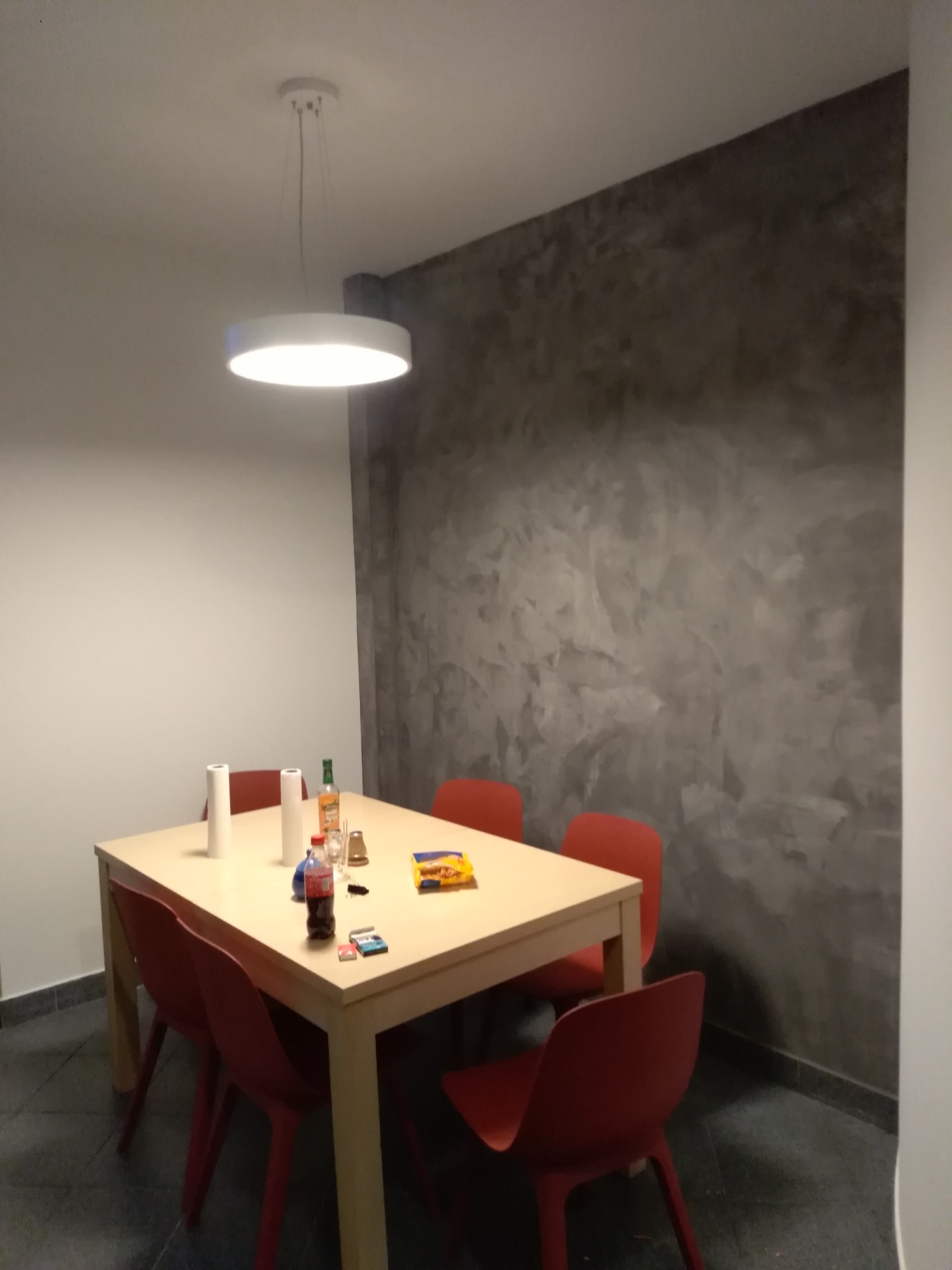Jadalnia po remoncie: kremowy stół z czerwonymi krzesłami, na blacie butelki, przekąski i rolka papieru, szara ściana z efektem betonu w tle, oświetlenie sufitowe LED.