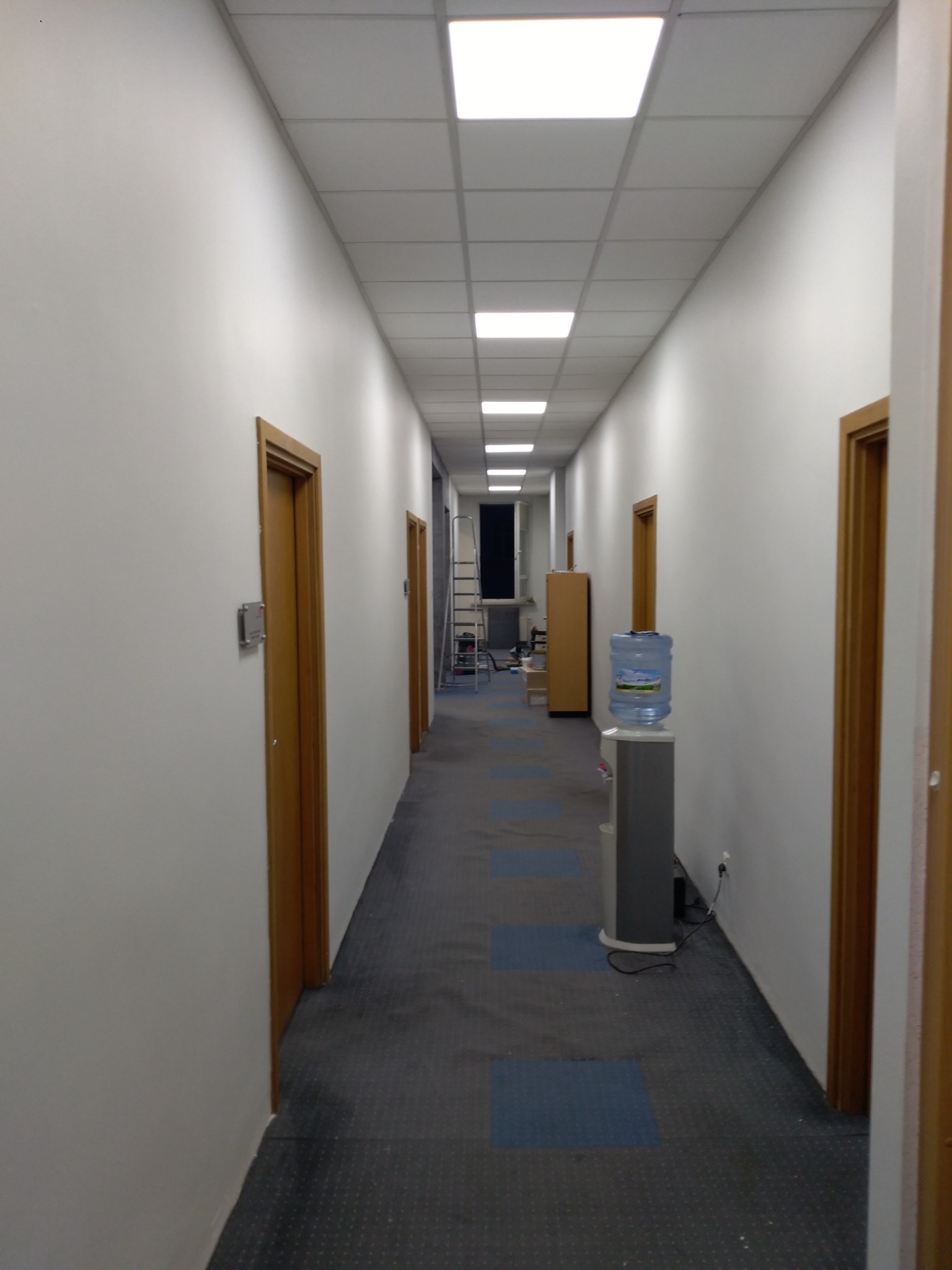 Długi korytarz biurowy z białymi ścianami, drewnianymi drzwiami, wykładziną w odcieniach szarości i niebieskich kwadratów, widoczne oświetlenie sufitowe i dystrybutor wody.