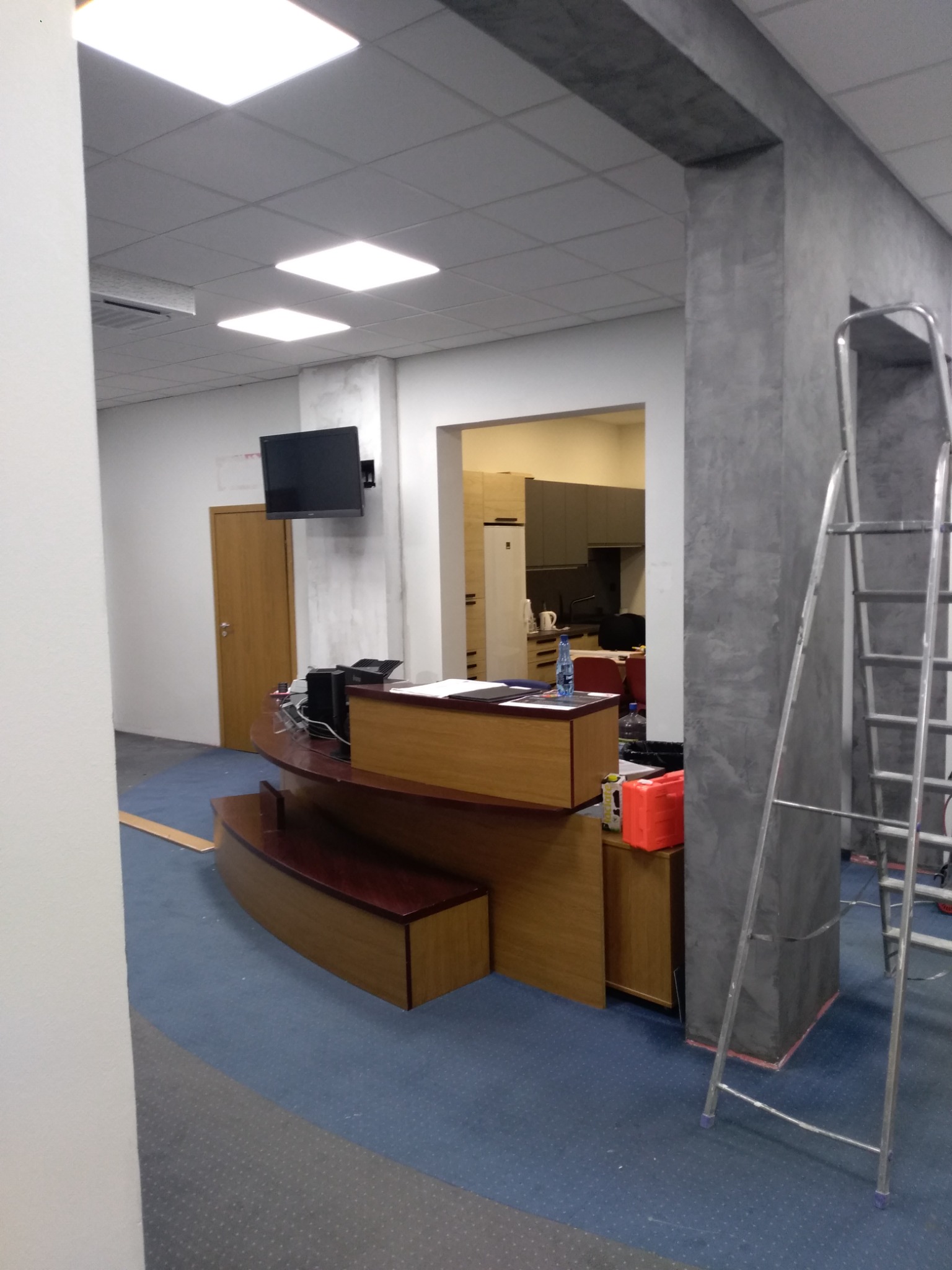 Biuro po remoncie: drewniana lada recepcyjna z komputerem, telewizor na ścianie, szara kolumna dekoracyjna, aluminiowa drabina, niebieska wykładzina dywanowa.