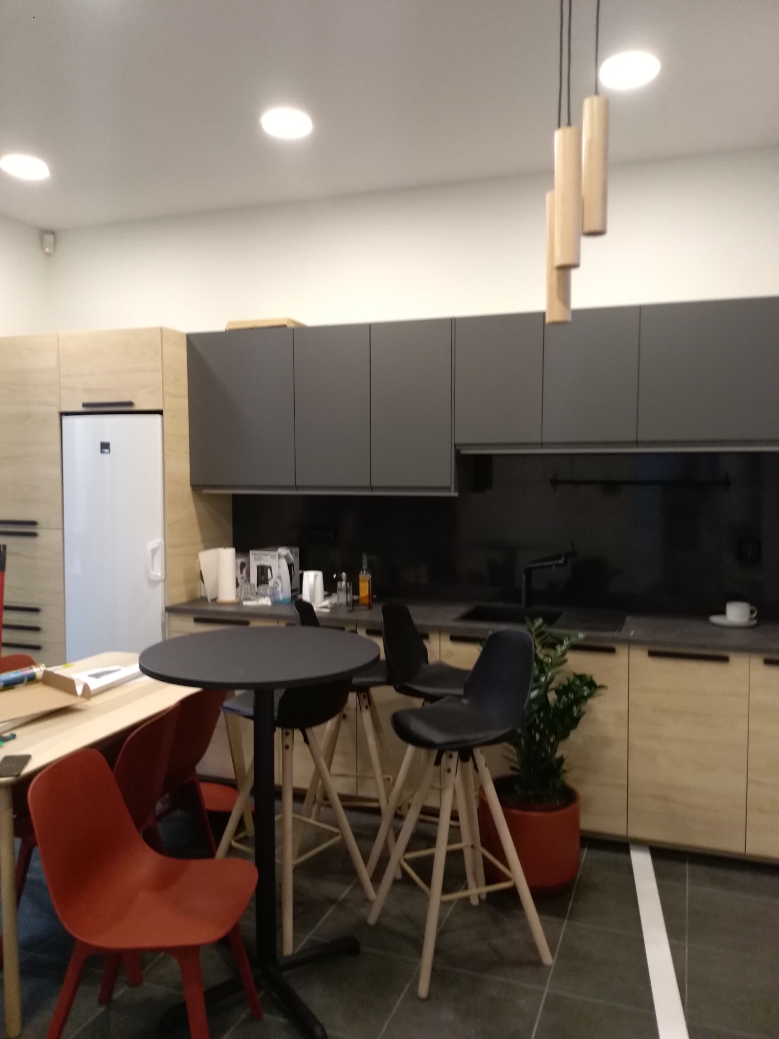 Nowoczesna kuchnia z szarymi szafkami górnymi i drewnopodobnymi dolnymi, czarnym blatem i stołem barowym z krzesłami, oświetlona trzema cylindrycznymi lampami wiszącymi, z widoczną rośliną...