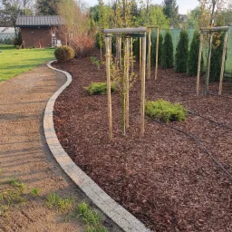 Krawężnik z kostki brukowej oddzielający ścieżkę od rabaty z korą w ogrodzie w Warszawie, widoczne młode drzewka i system nawadniania.