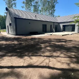 Nowoczesny dom z szarą elewacją i grafitową dachówką, widoczny taras w trakcie budowy, przygotowany teren pod trawnik, na tyłach zaparkowany sprzęt budowlany.