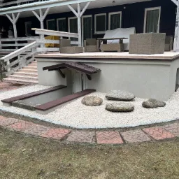 Wykończony taras na podwyższeniu z meblami ogrodowymi, obok wejście do piwnicy z ozdobnym kamieniem i obrzeżem z kostki.