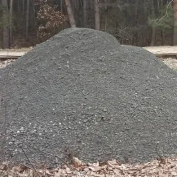 Duża pryzma szarego gruzu na tle lasu i tor&oacute;w kolejowych, widoczne suche liście i gałęzie na pierwszym planie.