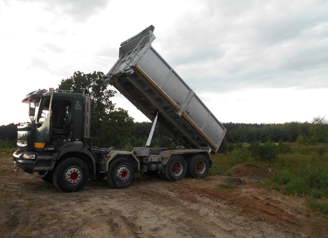 Ciężarówka z podniesioną skrzynią ładunkową, wysypująca materiał na ziemię w terenie niezabudowanym, w tle drzewa i pochmurne niebo.
