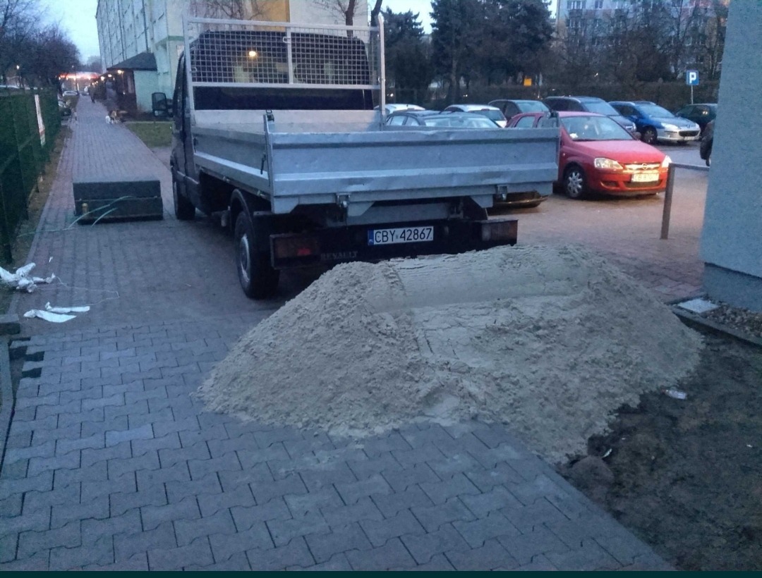 Samochód dostawczy z wywrotką zaparkowany na chodniku obok sterty piasku, w tle budynek mieszkalny i zaparkowane samochody.