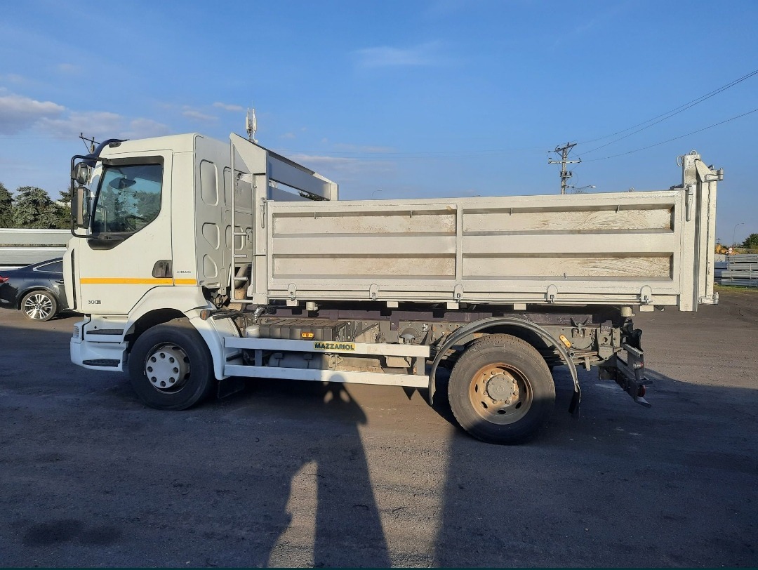 Biała wywrotka Renault 300 DXi z otwartą skrzynią ładunkową, zaparkowana na placu, widok z boku, cień osoby w kadrze.