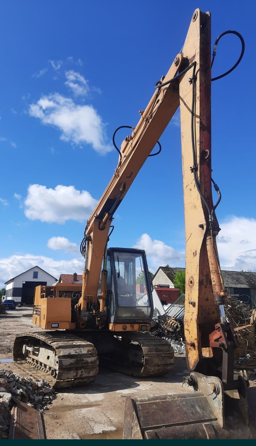 Żółta koparka gąsienicowa Poclain na placu budowy, fragmenty gruzu i złomu w tle, błękitne niebo z chmurami.