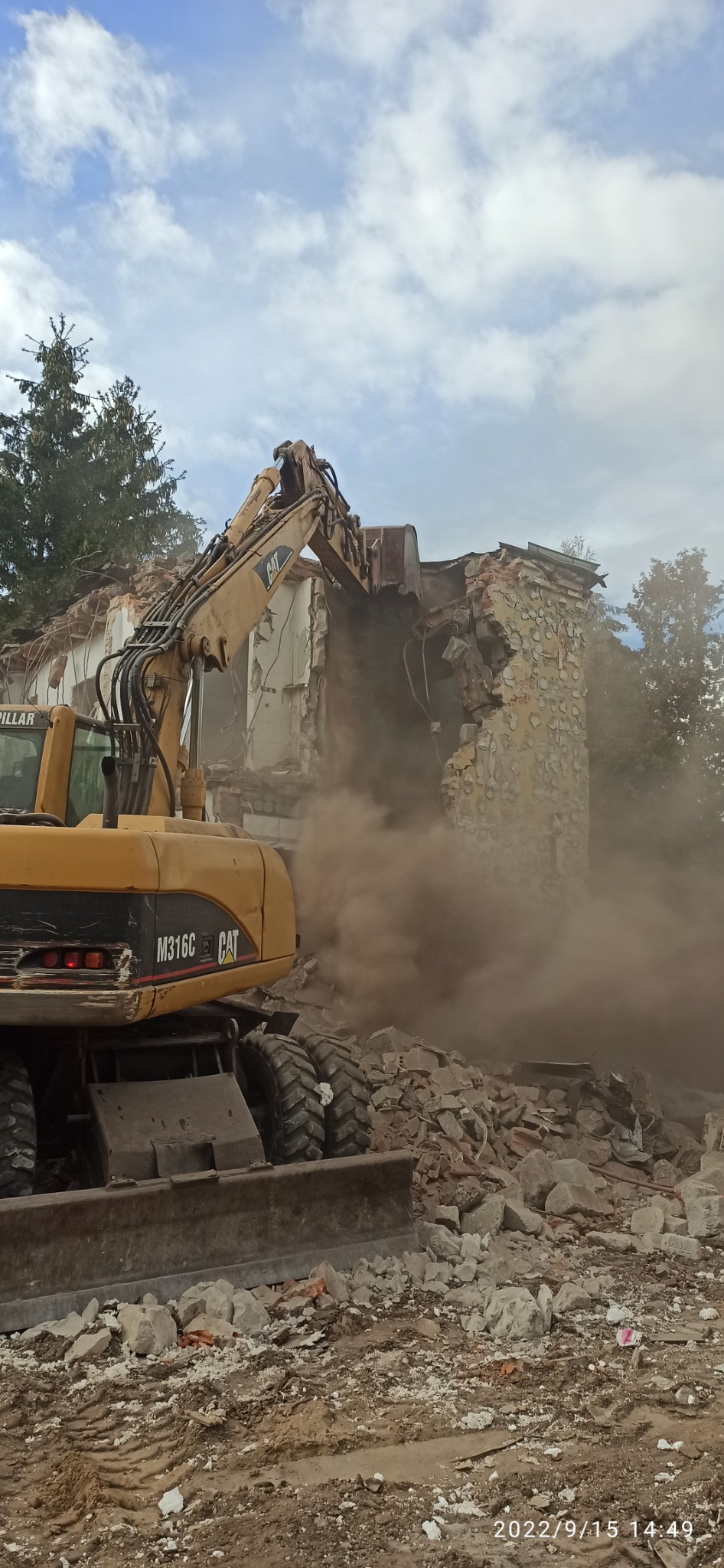 Żółta koparka CAT M316C w trakcie rozbiórki murowanego budynku, unoszący się pył i gruz.