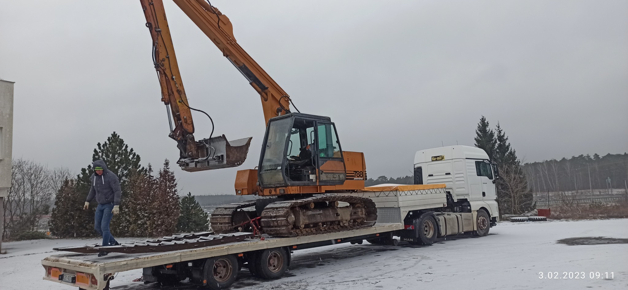 Pomarańczowa koparka gąsienicowa Poclain 788 POWERFIST załadowana na niskopodwoziową lawetę ciągniętą przez biały ciągnik siodłowy MAN, na śniegu, z operatorem w kabinie i mężczyzną w kurtce...