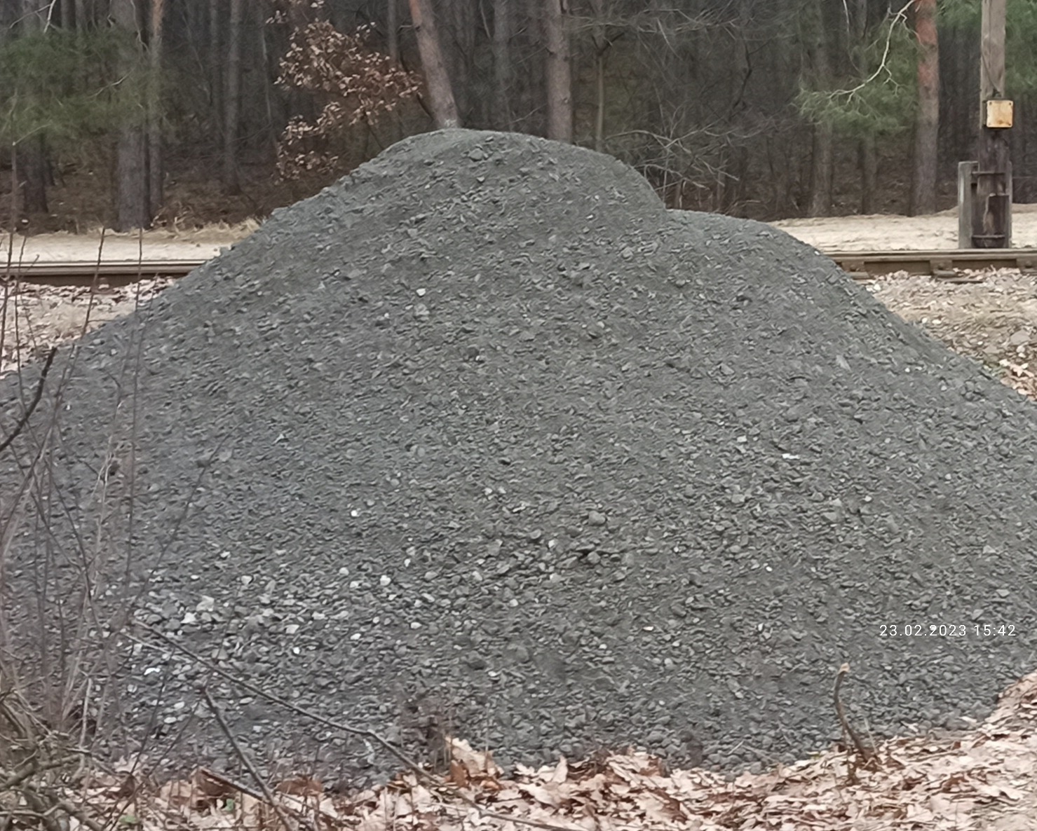 Duża pryzma szarego gruzu na tle lasu i torów kolejowych, widoczne suche liście i gałęzie na pierwszym planie.