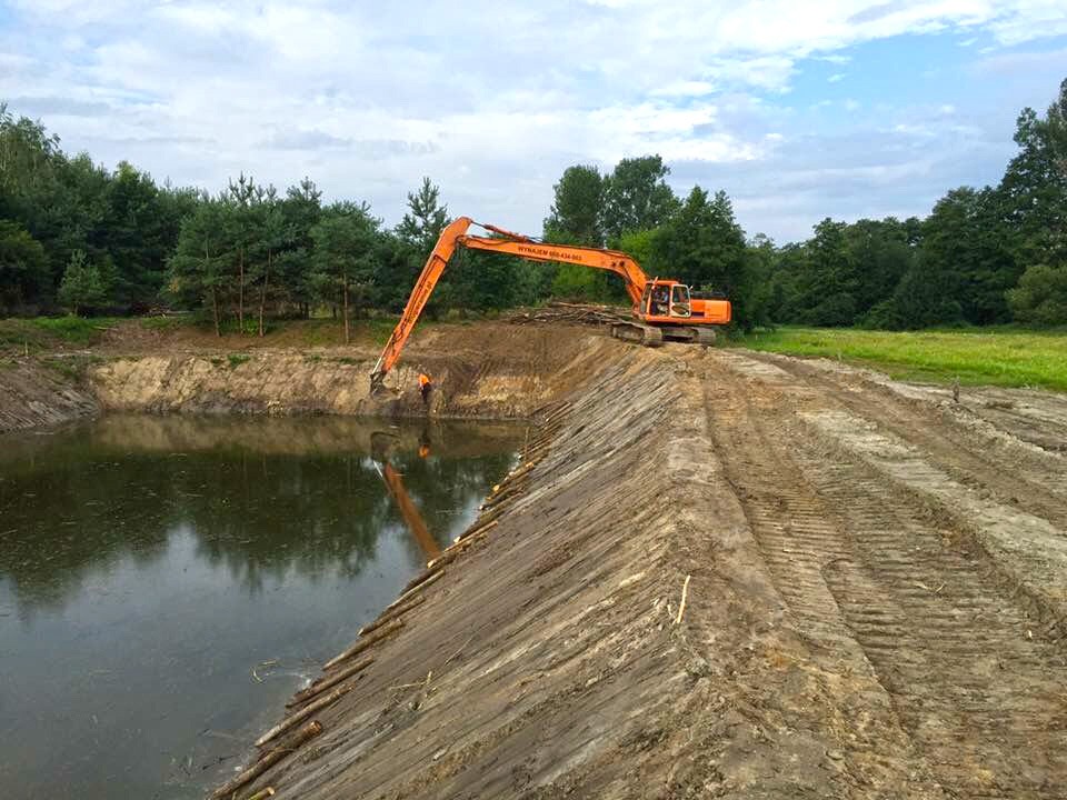Pomarańczowa koparka Hitachi 606 424 565 podczas pogłębiania brzegu nowo wykopanego stawu, widoczne ślady gąsienic na ziemi i pracownik w pomarańczowej kamizelce.