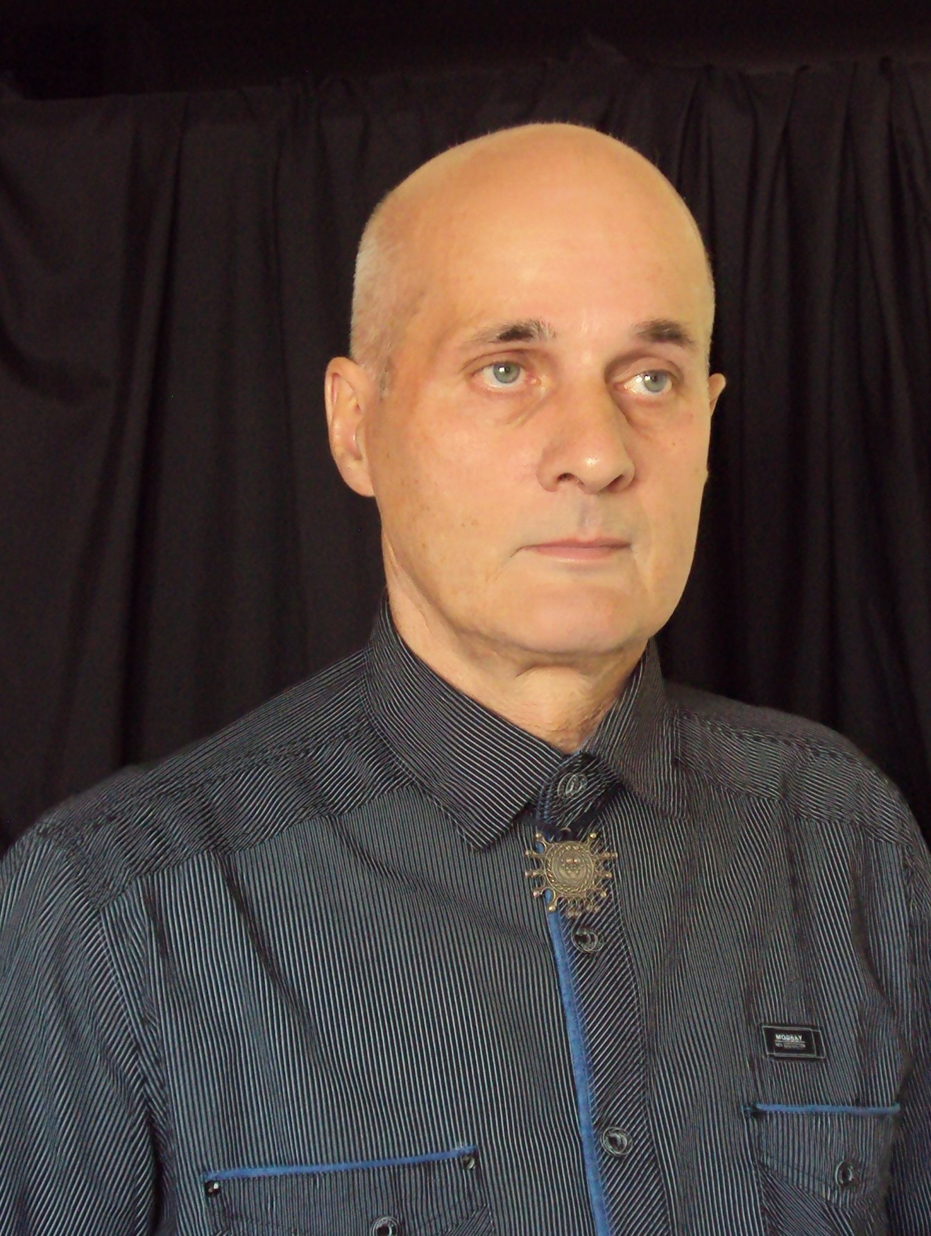 Portret mężczyzny z łysą głową w koszuli w paski z ozdobnym bolo tie na tle czarnej zasłony.