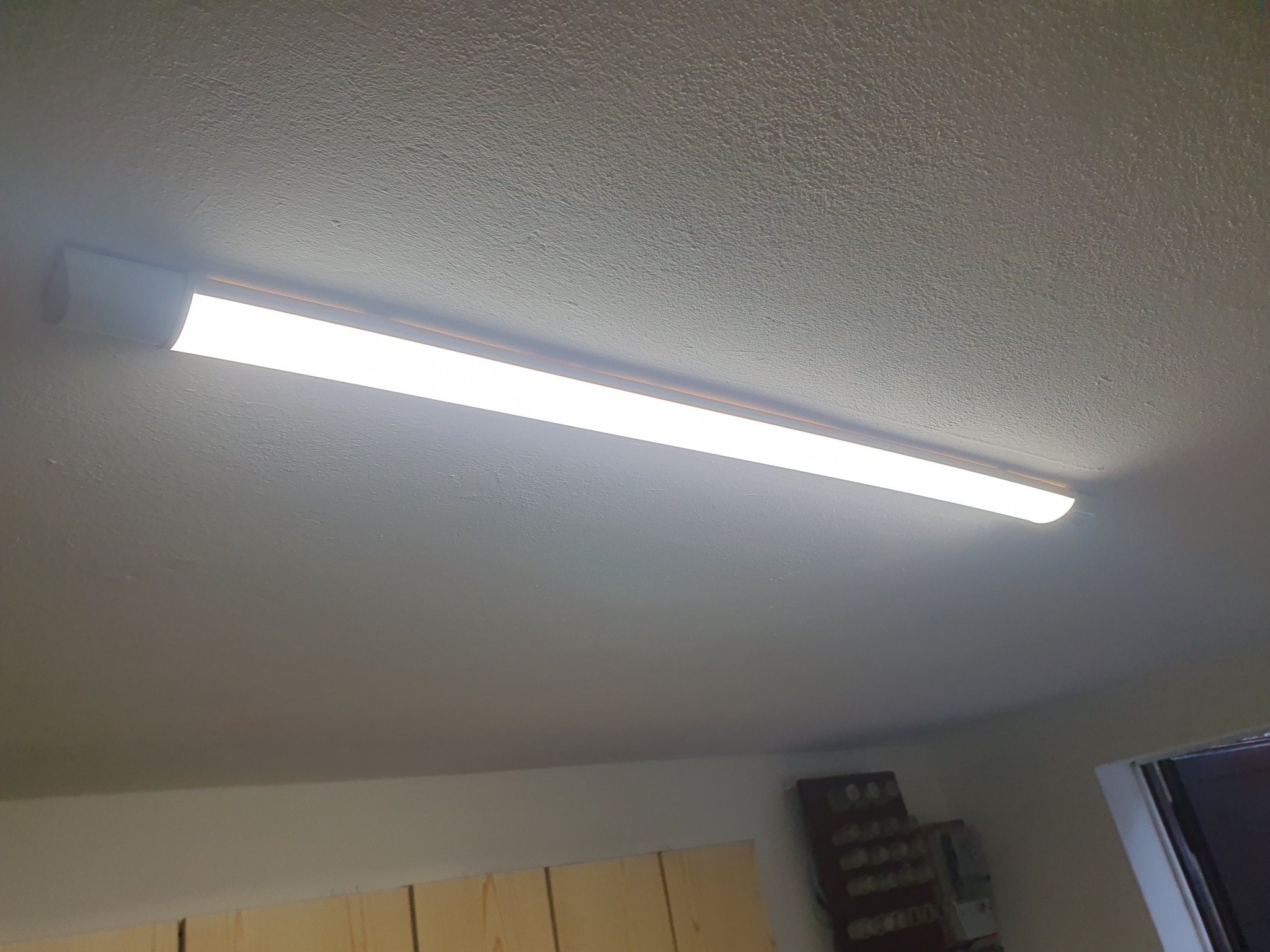 Zamontowana długa lampa LED na suficie w garażu, widoczna z dołu pod kątem, z fragmentem ściany i szafki w tle.