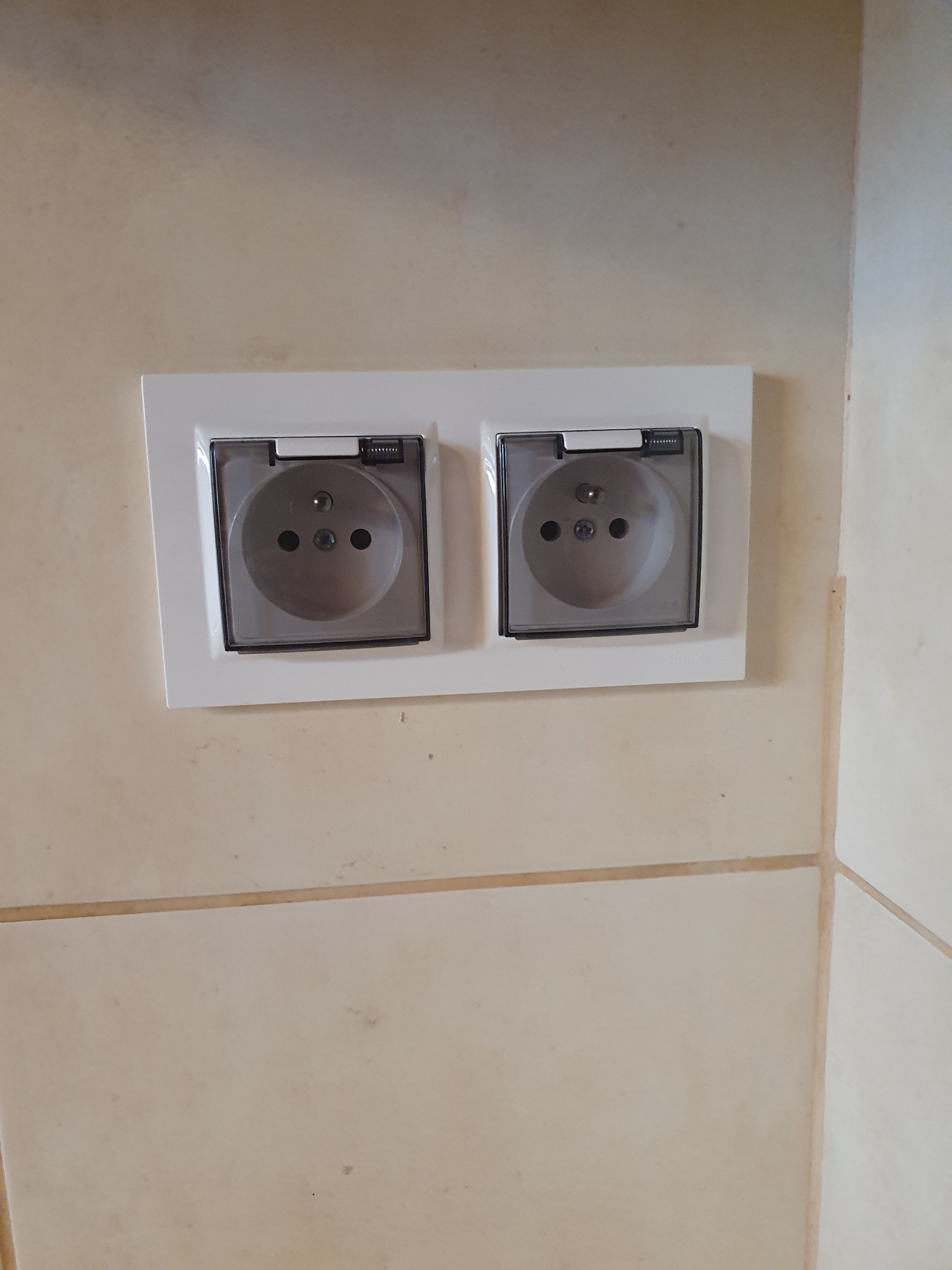 Podwójne gniazdko elektryczne z przezroczystymi klapkami ochronnymi zamontowane na jasnej, beżowej ścianie wyłożonej płytkami.