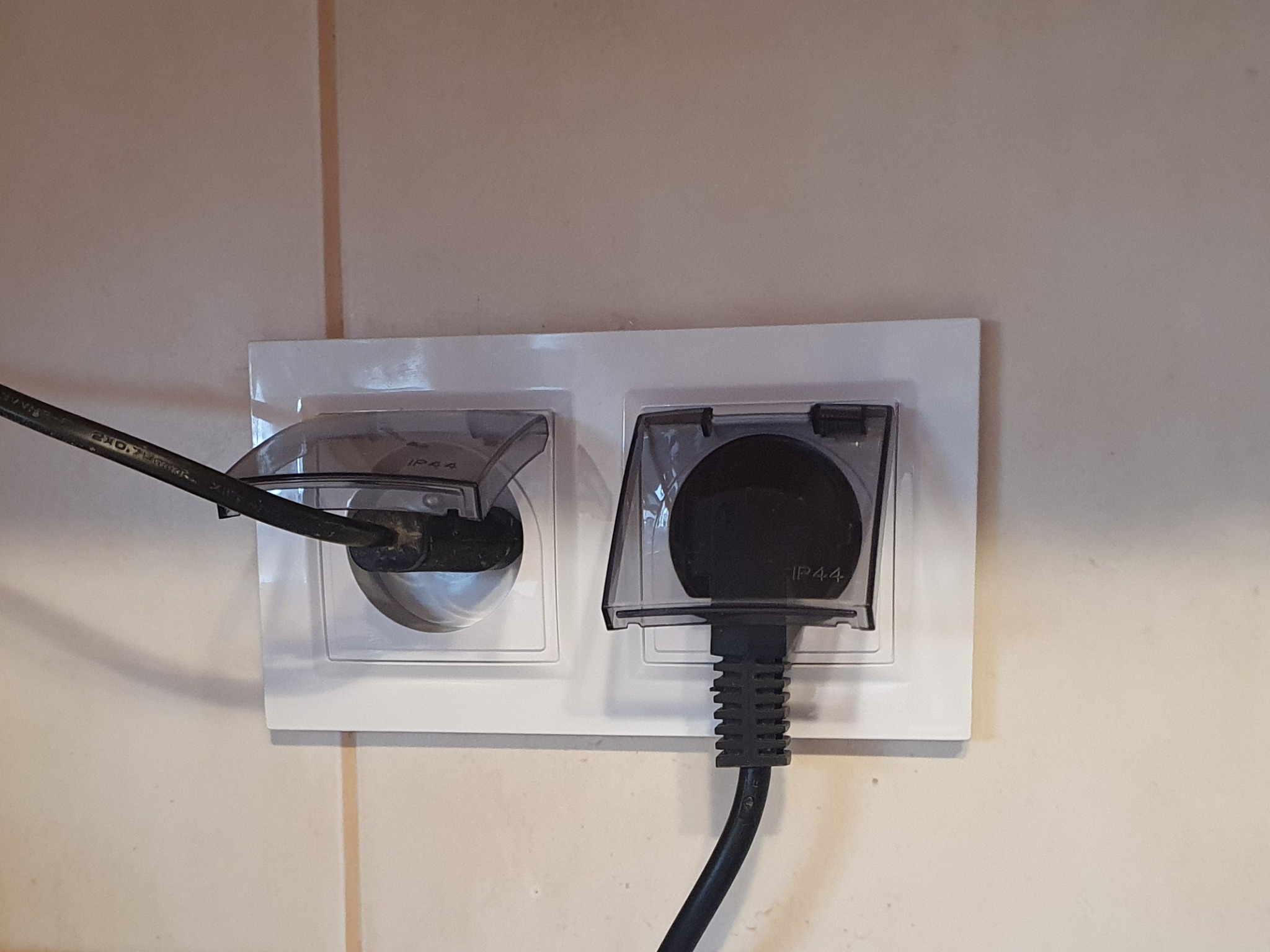 Podwójne gniazdko elektryczne z klapkami ochronnymi IP44, jedno z wpiętym przewodem zasilającym, zamontowane na kafelkowanej ścianie.