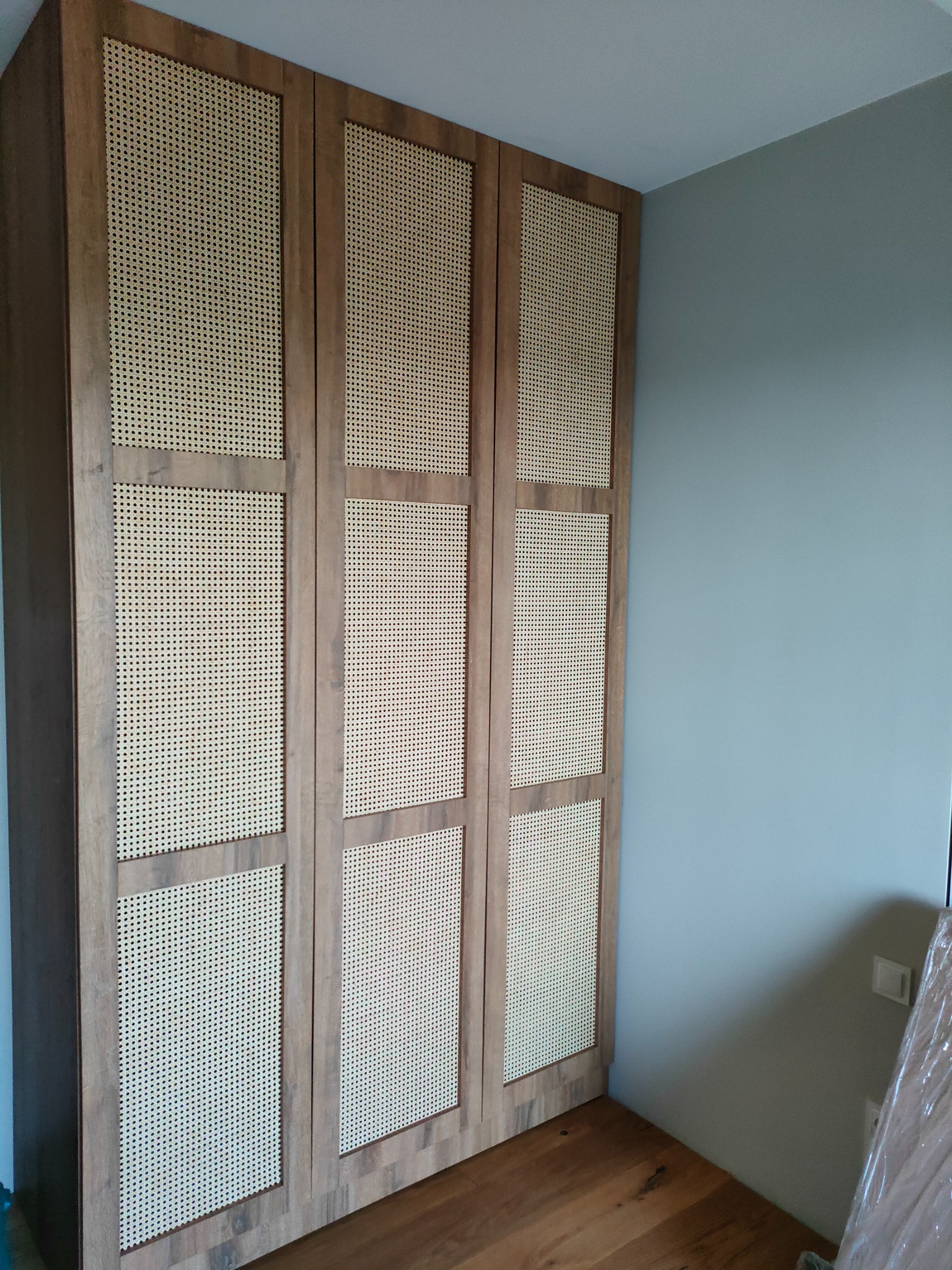 Trzydrzwiowa szafa z drewnianymi ramami i wypełnieniem z plecionki rattanowej, stojąca na podłodze z paneli, na tle szarej ściany.