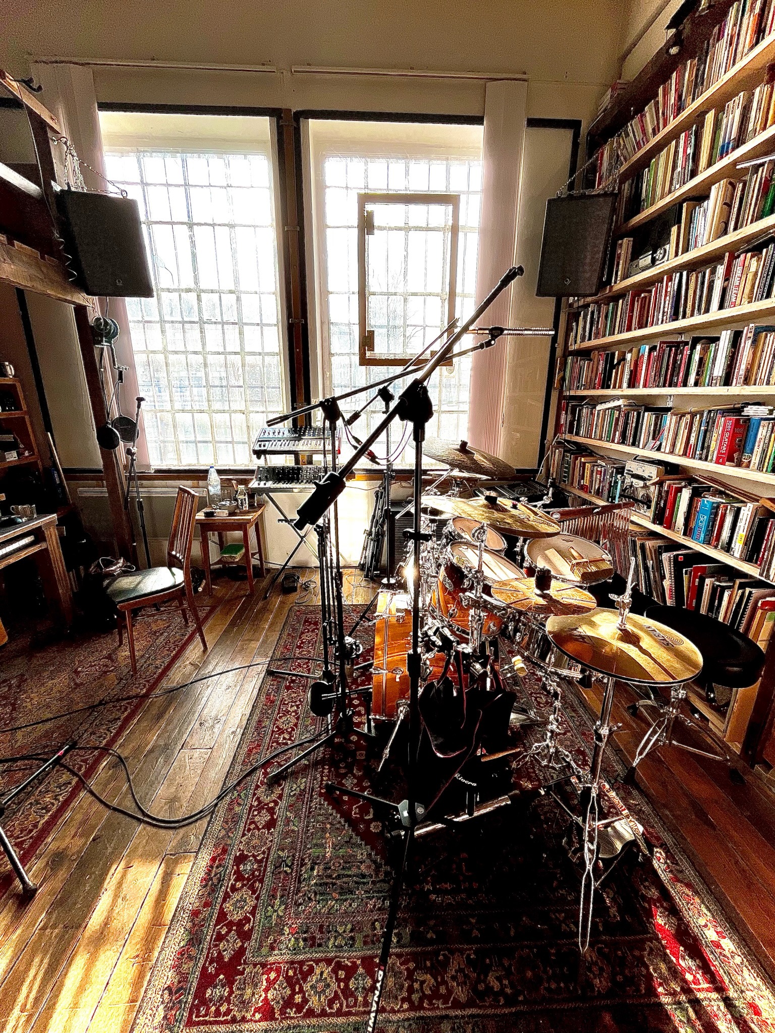 Perkusja ustawiona w pokoju z regałami pełnymi książek, oświetlona przez światło wpadające przez okno z kratownicą, widoczne mikrofony i keyboard.