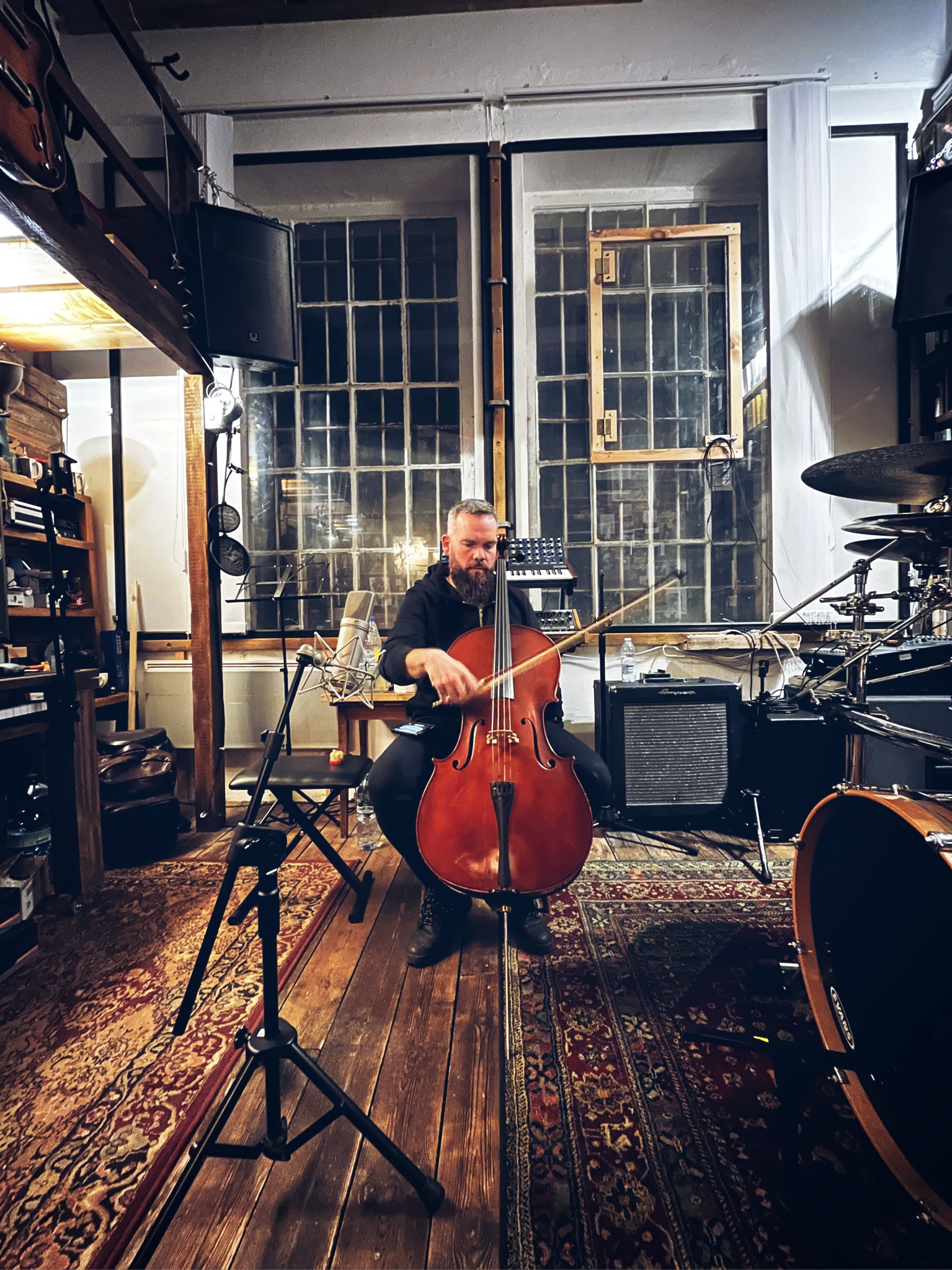 Muzyk z brodą gra na wiolonczeli w pomieszczeniu z instrumentami, mikrofonem i dużymi oknami w tle.