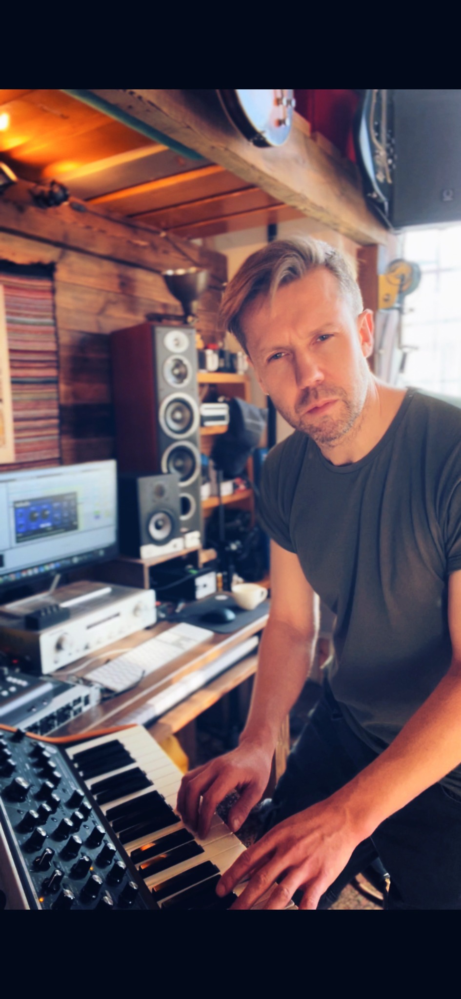 Mężczyzna grający na syntezatorze w domowym studiu nagrań z drewnianymi ścianami, widoczne monitory studyjne i ekran komputera z oprogramowaniem do edycji audio.