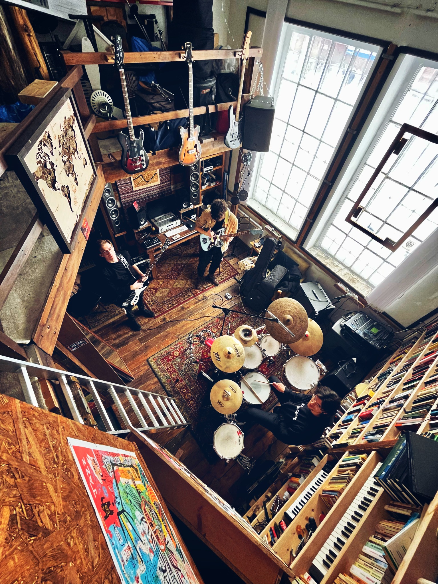 Skośny kadr z góry na muzyków grających w pomieszczeniu z oknami, instrumentami i książkami; gitary, perkusja, klawisze i mikser na dywanach orientalnych.