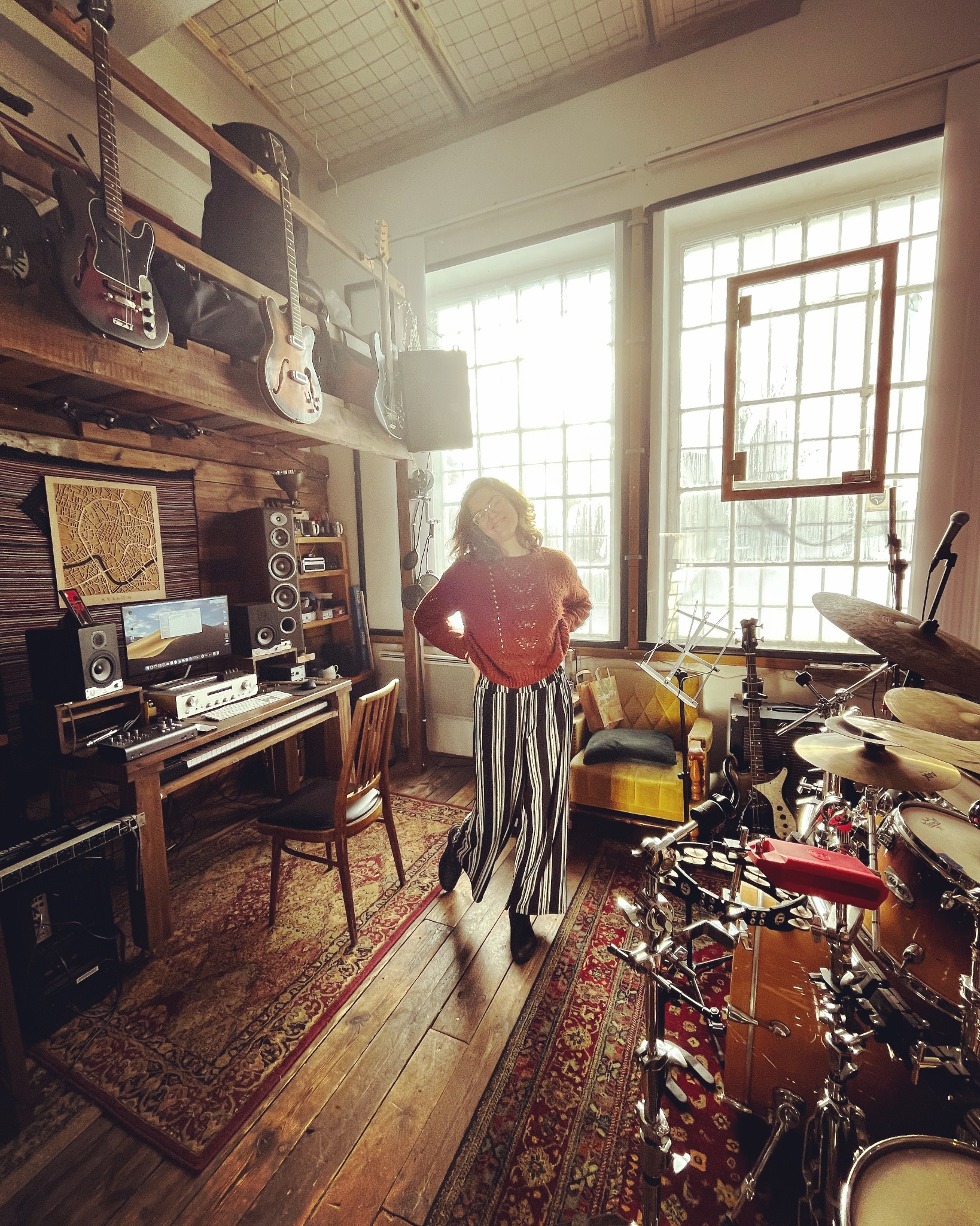 Kobieta w paskowanych spodniach stoi w przestronnym pokoju z instrumentami muzycznymi, w tle biurko z komputerem i głośnikami, gitary wiszące na półce pod sufitem, a w prawym dolnym rogu zestaw...