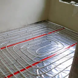 Białe rury ogrzewania podłogowego ułożone na folii odblaskowej z czerwoną siatką, przymocowane plastikowymi klipsami do podłoża w jasnym pomieszczeniu w trakcie montażu.