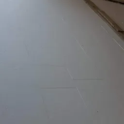 Białe płyty izolacyjne ułożone na podłodze, przygotowane pod montaż ogrzewania podłogowego, widoczne szczeliny między płytami i fragment okna z workami izolacji w tle.