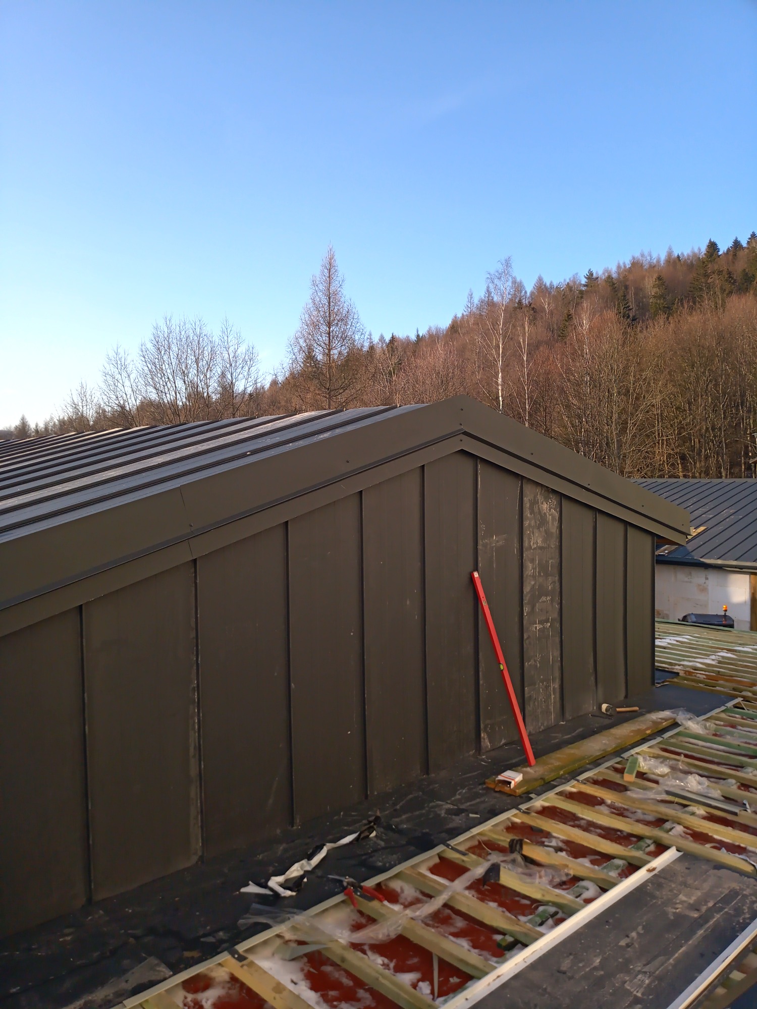 Montaż paneli na dachu budynku w trakcie prac, widoczny poziomica oparta o ścianę, elementy konstrukcji dachu i ocieplenie, w tle drzewa i błękitne niebo.