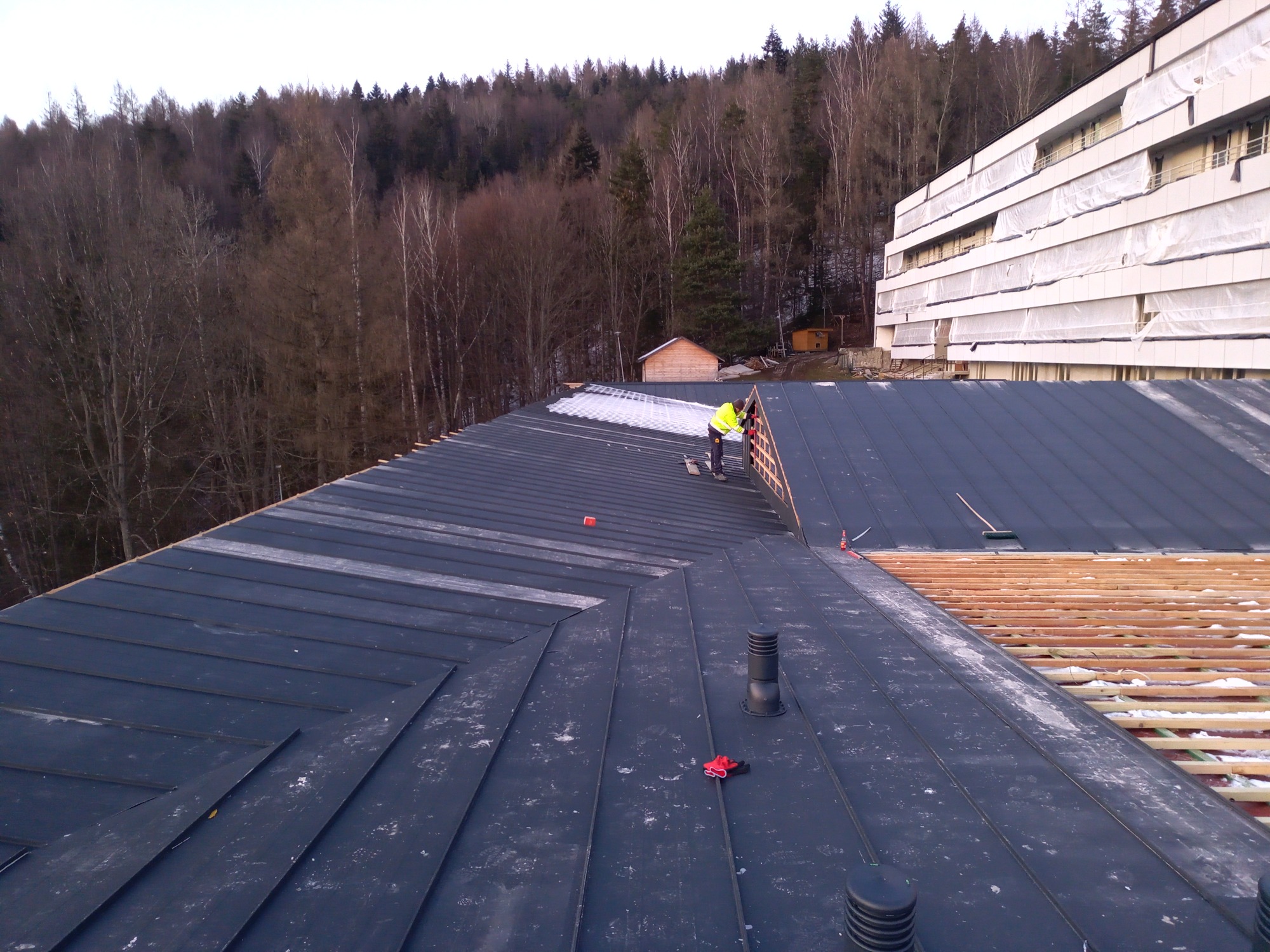 Prace dekarskie na dachu budynku wielorodzinnego, widok z góry na fragment pokrycia z blachy na rąbek stojący, częściowo ośnieżonego, z widoczną więźbą dachową w tle i dekarzem w żółtej kamizelce...