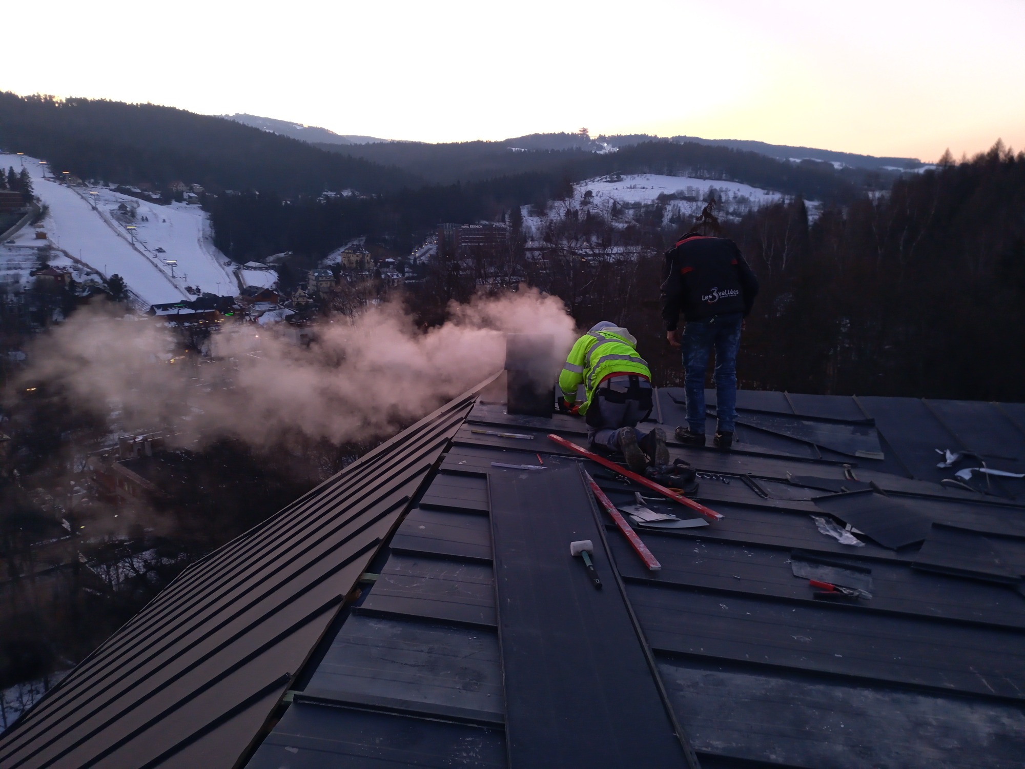 Dwóch dekarzy pracuje na dachu krytym blachą na rąbek stojący, w tle widok na ośnieżone wzgórza i dym z komina.