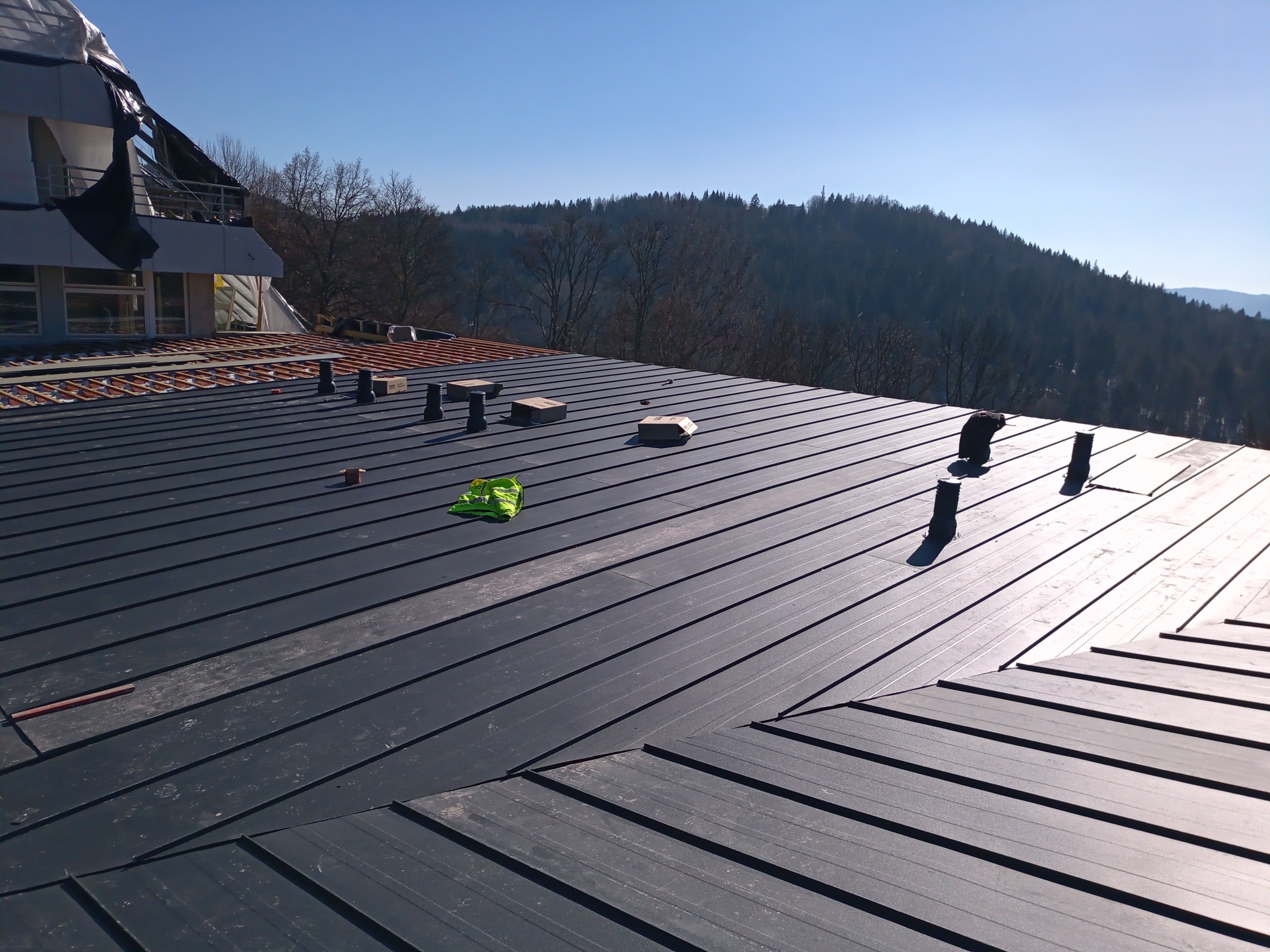 Nowoczesny dach z blachy na tle zalesionych wzgórz, widoczny fragment budynku z osłoniętym rusztowaniem, elementy wentylacyjne i robocza kamizelka odblaskowa.