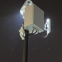 Maciej Wegiera ELEKTRONIK - Zewnętrzna kamera monitoringu z reflektorem na słupie, widoczna w nocy, z widocznymi kablami i puszką przyłączeniową.