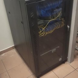 Maciej Wegiera ELEKTRONIK - Czarna szafa rack z przeszklonymi drzwiami, wypełniona niebieskimi i żółtymi kablami sieciowymi, stojąca na kafelkach pod ścianą.