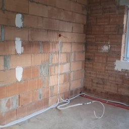 Maciej Wegiera ELEKTRONIK - Surowy mur z czerwonej cegły z widocznymi przewodami elektrycznymi w białych rurkach i puszkami instalacyjnymi w kolorze pomarańczowym; widoczne także rury wodne i okno z folią ochronną.