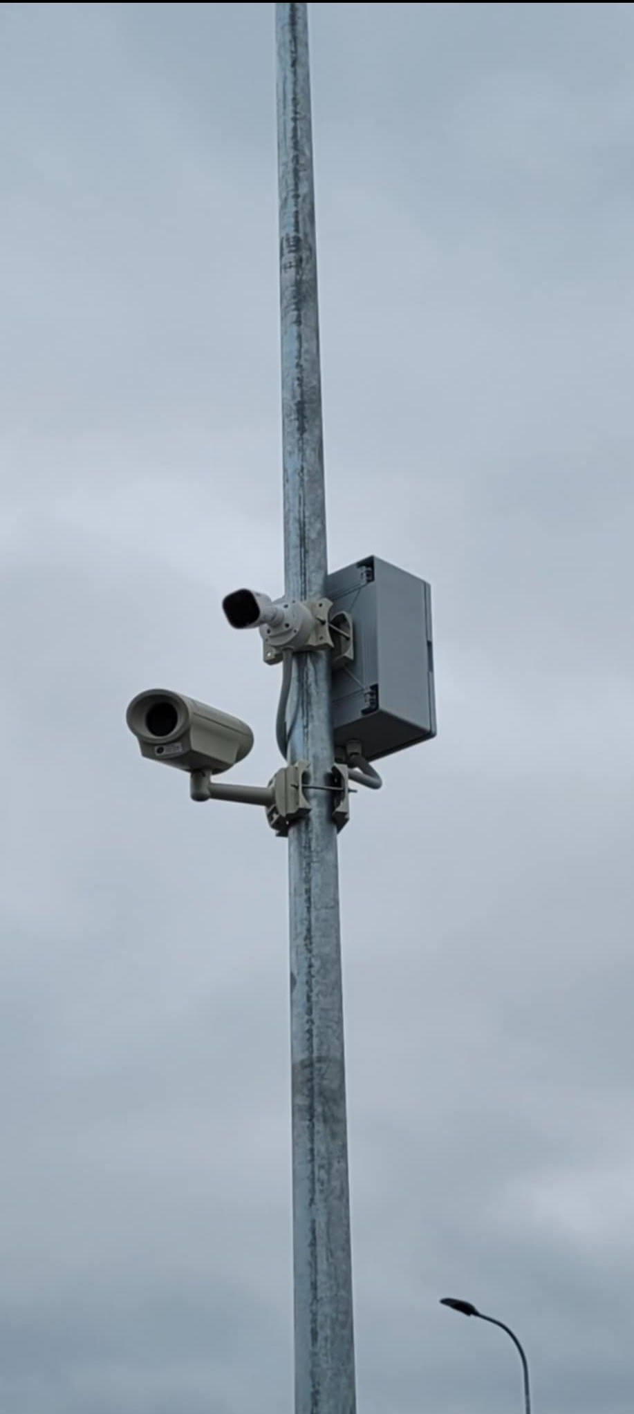 Dwie kamery przemysłowe zamontowane na metalowym słupie, szara obudowa z elektroniką, pochmurne niebo w tle.