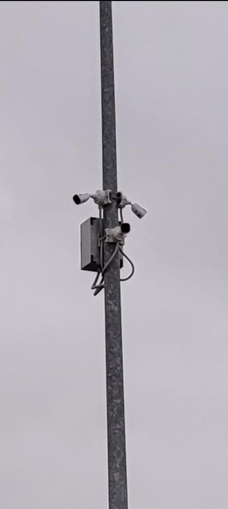 Słup z trzema kamerami przemysłowymi, szarą skrzynką i przewodami, widoczne na tle pochmurnego nieba.