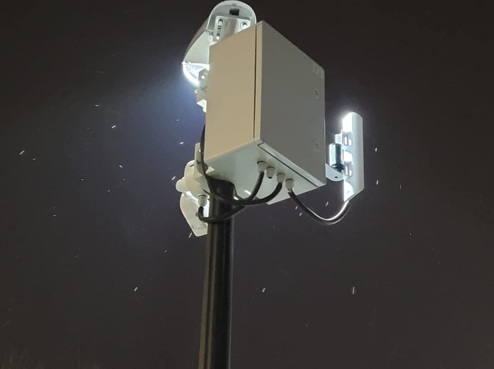 Zewnętrzna kamera monitoringu z reflektorem na słupie, widoczna w nocy, z widocznymi kablami i puszką przyłączeniową.