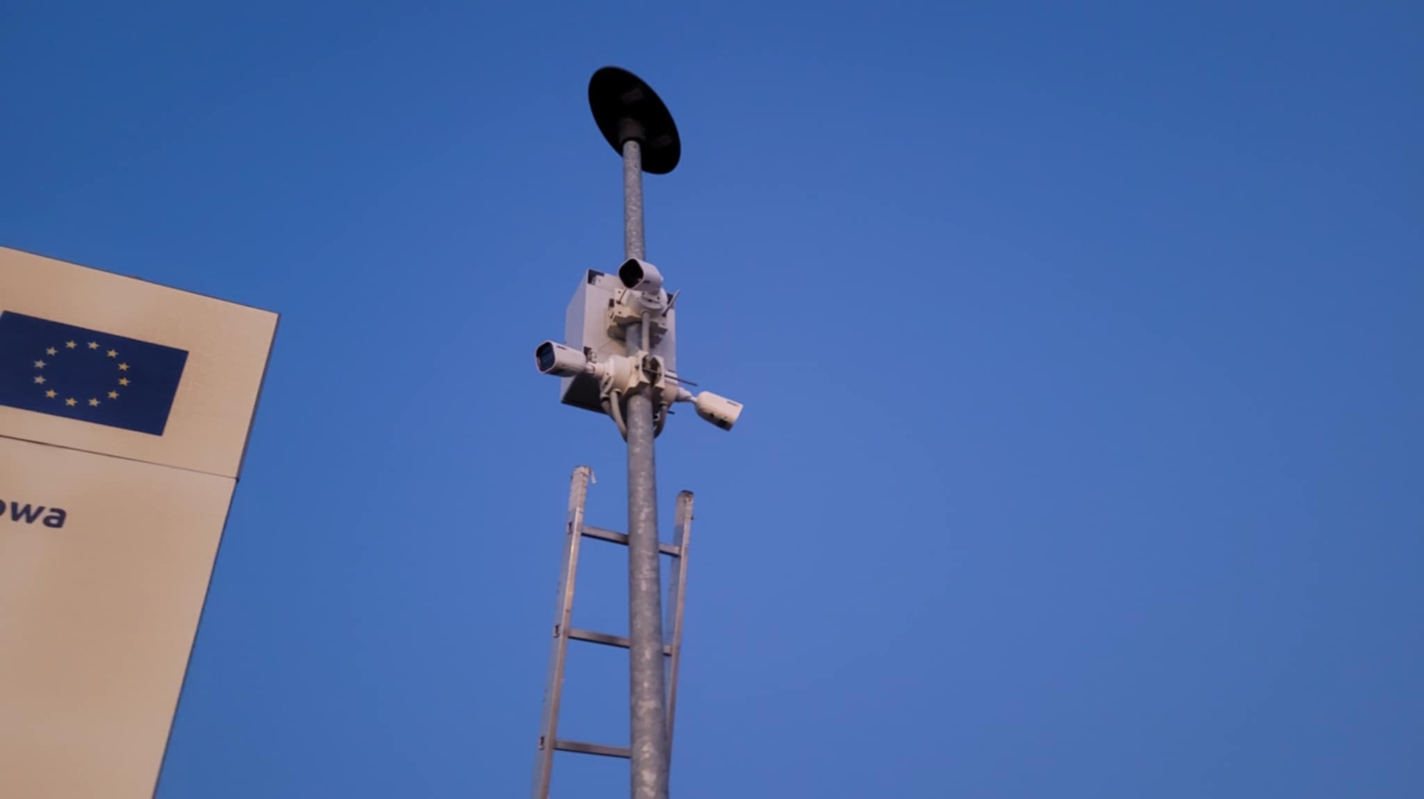 Słup z zamontowanymi kamerami przemysłowymi i drabiną na tle błękitnego nieba, obok widoczny znak z flagą Unii Europejskiej.