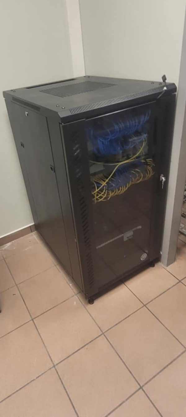 Czarna szafa rack z przeszklonymi drzwiami, wypełniona niebieskimi i żółtymi kablami sieciowymi, stojąca na kafelkach pod ścianą.